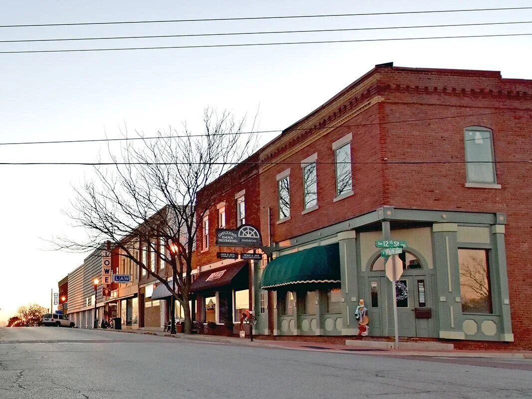 Two-story historic brick storefronts with a green awning at the corner of Main and 12th Street at dusk, with bare trees along the sidewalk