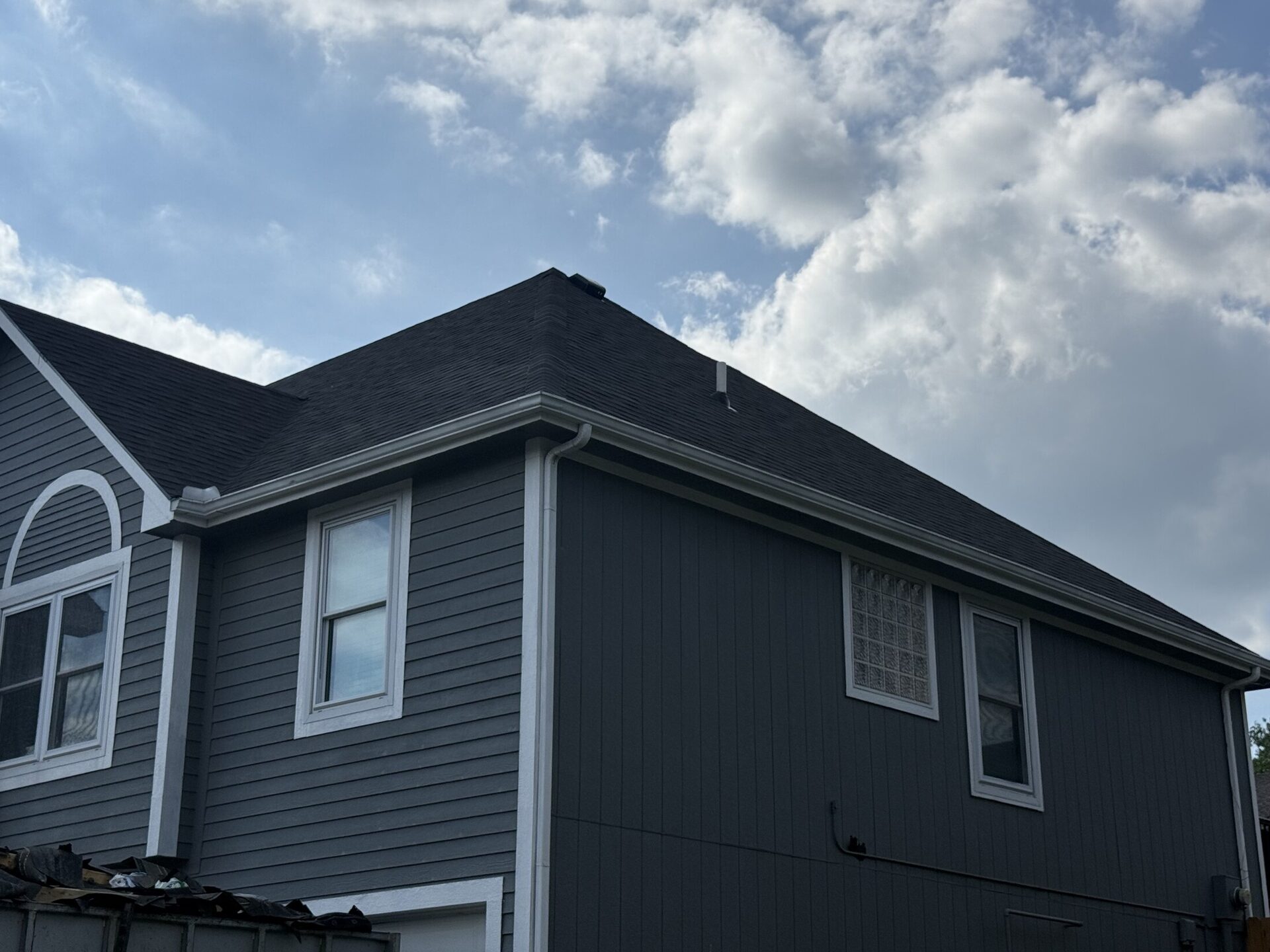A gray two-story home with a new charcoal shingle roof and white gutters under a cloudy sky, with a dumpster of torn-off shingles in front