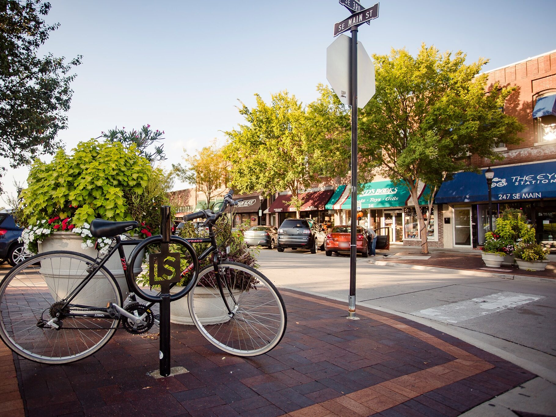 A bicycle locked to an LS bike rack at the corner of SE Main St and SE 3rd St, with awninged brick storefronts and parked cars along the street