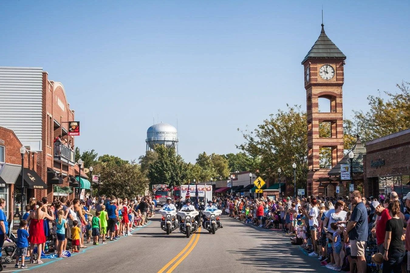 A parade passing the brick clock tower in downtown Overland Park, with the Overland Park water tower behind it