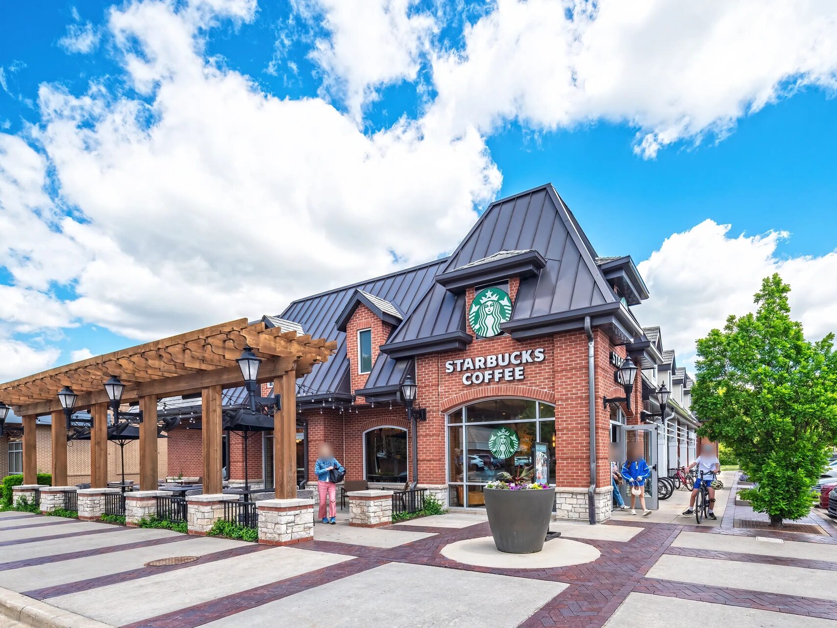A red-brick Starbucks with a dark standing-seam metal roof, dormers and a wooden pergola at a shopping center, under a bright blue sky with white clouds